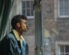 Young man in a denim jacket pauses by a window, looking outside