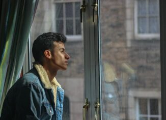 Young man in a denim jacket pauses by a window, looking outside